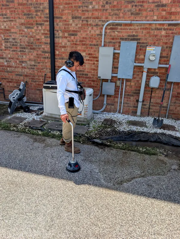 Technician scanning for a hidden leak beside a brick home with electronic leak detection gear